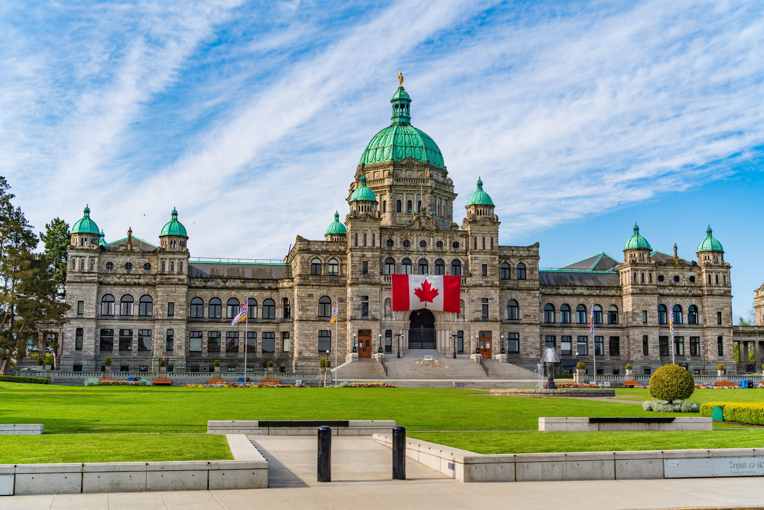 Front view of a grand stone civic building with a large green dome, smaller domes, and a Canadian flag flying above the entrance set against a blue sky.