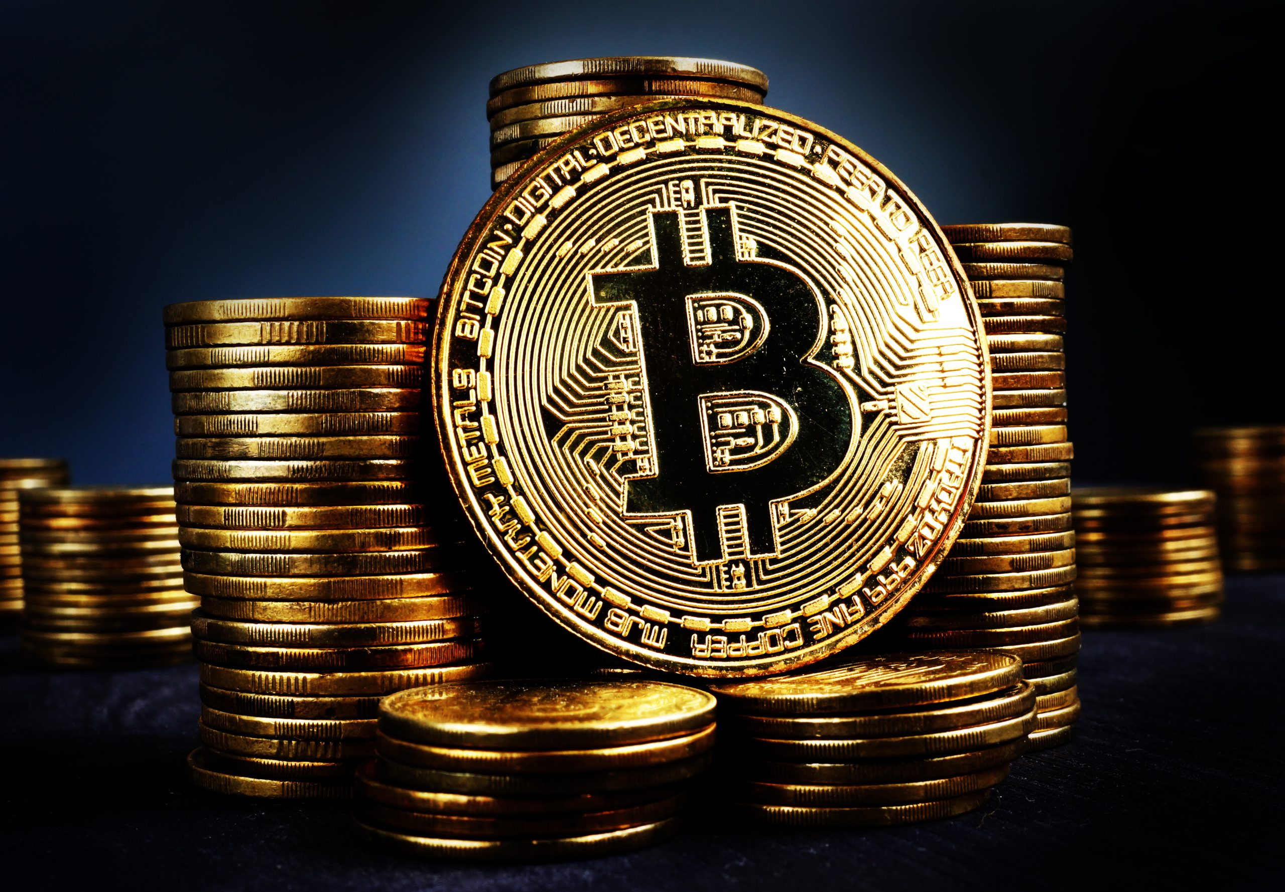 Stack of gold Bitcoin coins with the Bitcoin symbol in the foreground on a dark background.
