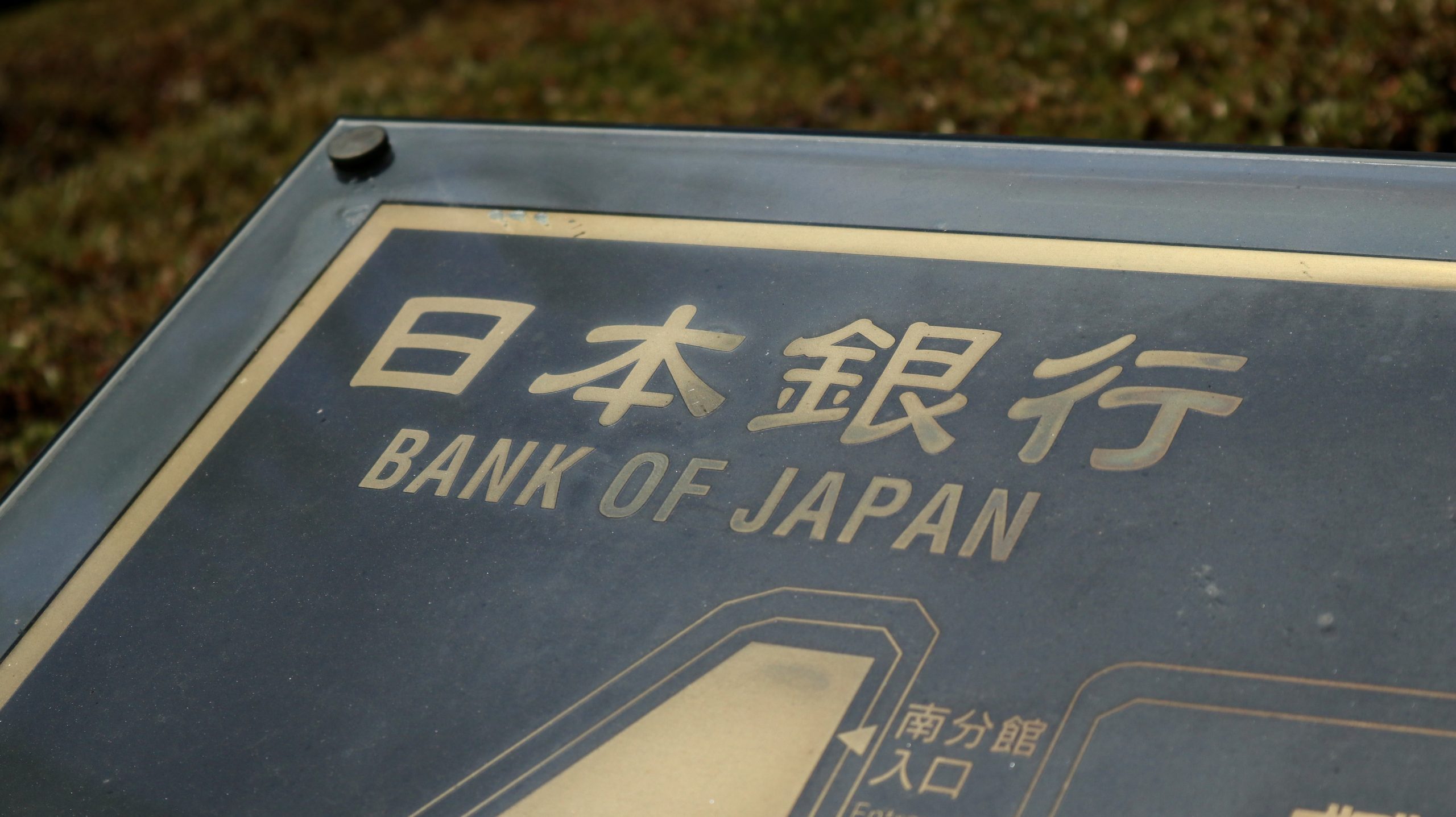 Close-up of an outdoor sign reading Bank of Japan in English with Japanese characters, shown at an angle on a metal frame.