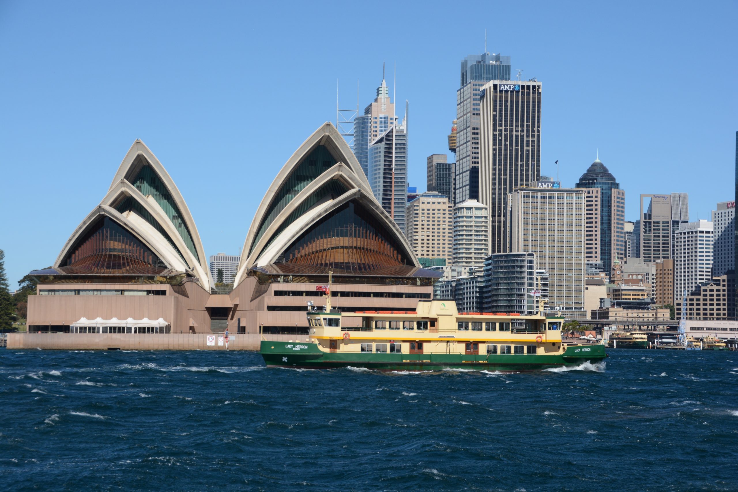 Sydney Opera House sails in the foreground with the city skyline behind and a green-and-yellow ferry on blue water in the foreground.