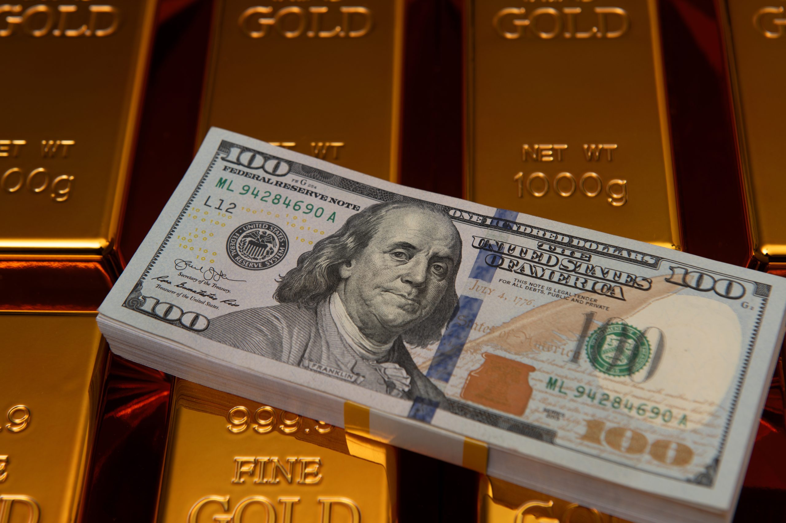 Stack of gold bars with a $100 bill laid across the foreground, hinting at wealth and precious metal value.