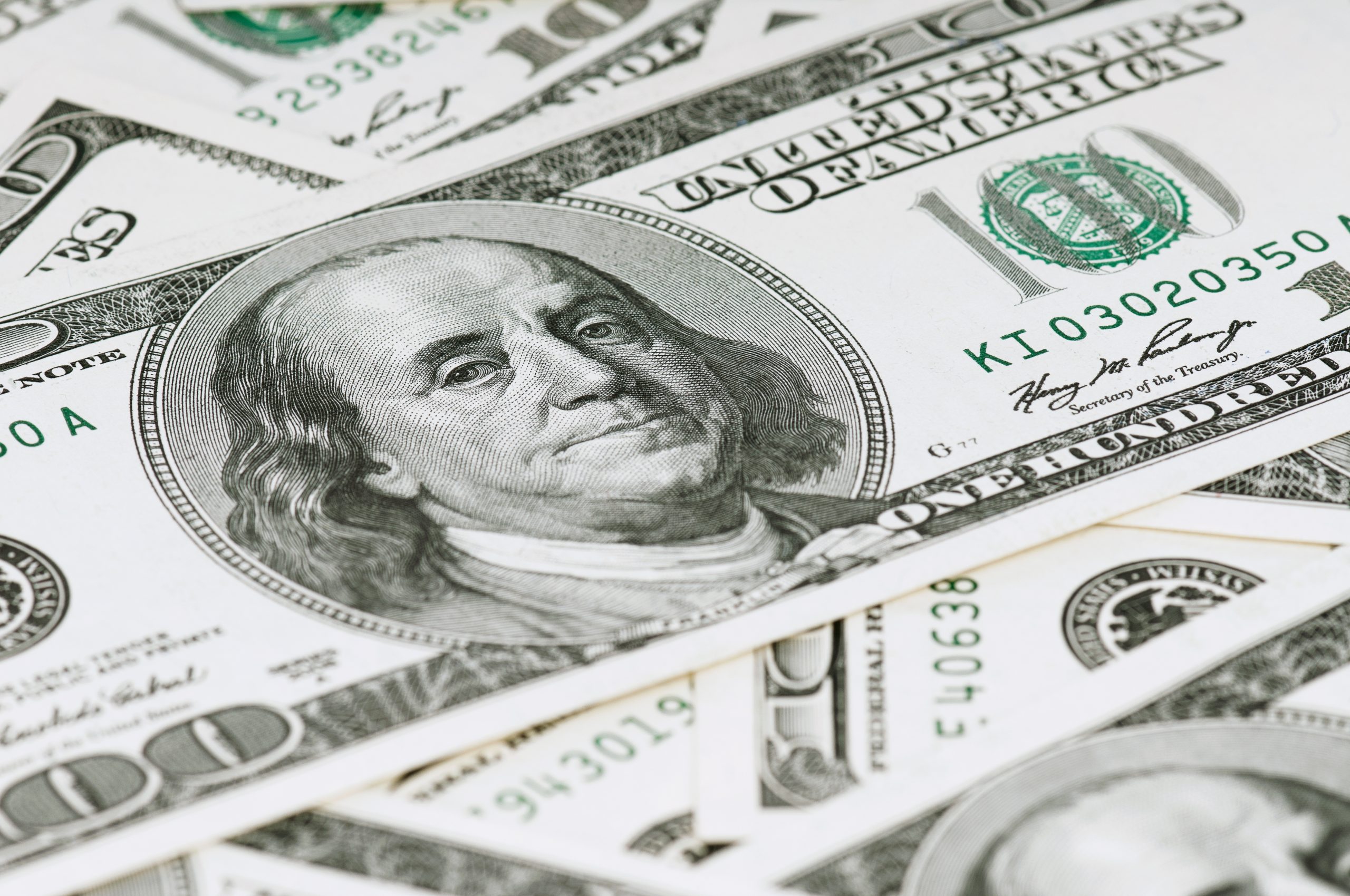 Close-up of overlapping U.S. hundred-dollar bills featuring Benjamin Franklin, with green ink and intricate engravings visible across the stack.