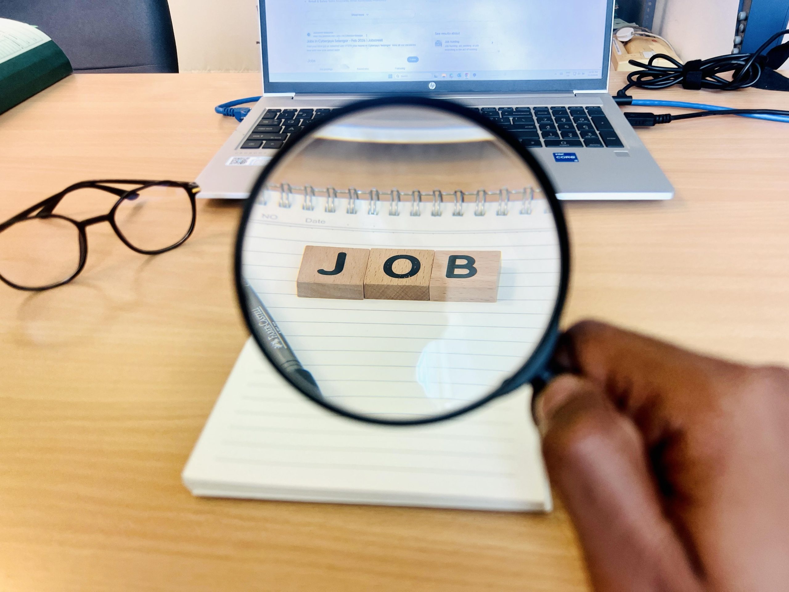 Magnifying glass over a notebook with wooden letters J, O, B on a desk; laptop, glasses, and cables in the background.